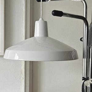 May include: A white, modern pendant light with a wide, conical shade. The light fixture is suspended from a metal frame and has a clean, minimalist design. The shade is approximately 18 inches in diameter.