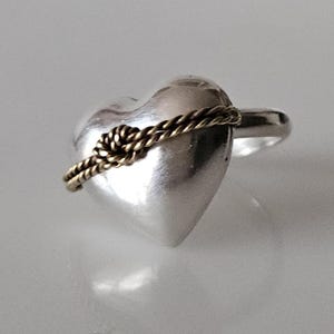 Op de afbeelding: Een zilveren ring in de vorm van een hart met een gouden touw eromheen gewikkeld.