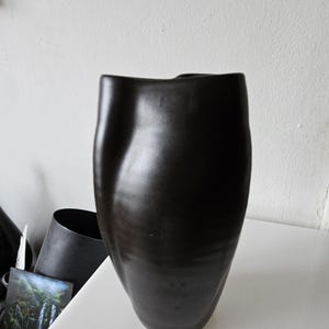 May include: A tall, matte black ceramic vase with a smooth, organic, and asymmetrical silhouette. The vase features a curved, undulating body that tapers toward the base and has a wide, irregular opening at the top, showcasing a solid dark finish.