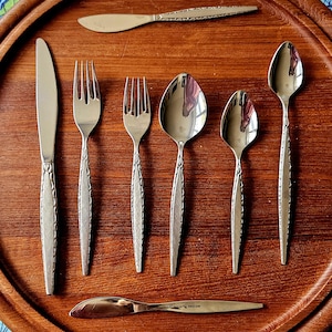 Puede incluir: Un conjunto de cubiertos de acero inoxidable, que incluye cuchillos, tenedores y cucharas, dispuestos sobre una bandeja de madera. La cubertería tiene un diseño decorativo en los mangos. La bandeja es de color marrón cálido.