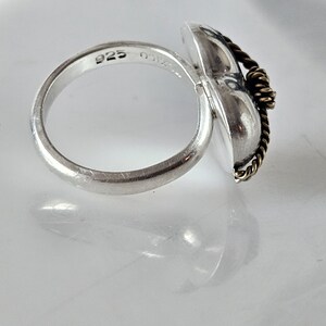 Op de afbeelding: Een zilveren ring met een hartvormig ontwerp en een gedraaid gouden draadaccent. De ring is gemarkeerd met "925" aan de binnenkant van de band.