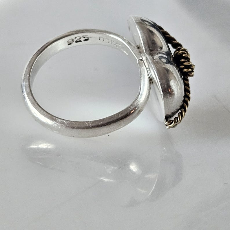 Op de afbeelding: Een zilveren ring met een hartvormig ontwerp en een gedraaid gouden draadaccent. De ring is gemarkeerd met "925" aan de binnenkant van de band.
