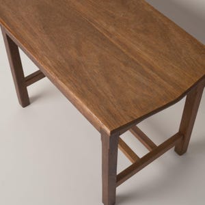 May include: A small wooden stool with a rectangular top and four straight legs connected by horizontal support bars. The stool features a natural medium brown wood grain finish with a smooth surface and visible wood texture throughout the piece.