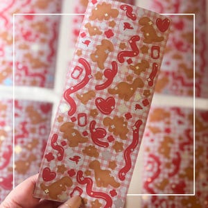 Puede incluir: Una hoja de pegatinas con un diseño de cuadros rosas y varias ilustraciones. Las pegatinas incluyen formas de dinosaurios marrones, corazones rojos, líneas onduladas y pequeñas formas de nubes y flores blancas. Las pegatinas son sostenidas por una mano.