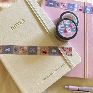 May include: A cream-colored notebook with the word NOTES and THOUGHTS, REFLECTIONS & IDEAS embossed in gold, a pink notebook, a roll of washi tape with a patchwork pattern of cats and cherries, and a light pink pen with a matching tape strip.