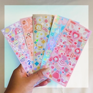 Puede incluir: Una mano sostiene cinco hojas de pegatinas con varios diseños en colores pastel. Las pegatinas presentan ilustraciones de comida, animales y elementos de circo. Las hojas tienen un acabado holográfico y brillante, añadiendo un efecto brillante a los diseños.
