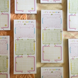 May include: A collection of rectangular paper notepads featuring a grid pattern of shopping baskets in pink, yellow, and blue. Each notepad displays the text LET'S GO SHOPPING at the top, accompanied by small star and heart icons.