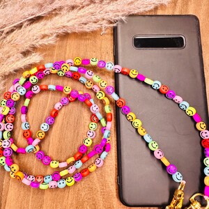 Könnte beinhalten: Ein farbenfroher Handy-Anhänger aus kleinen Smiley-Perlen. Der Anhänger ist an einer schwarzen Handyhülle befestigt und hat einen goldenen Metallverschluss.