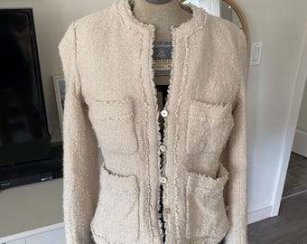 May include: A beige tweed jacket featuring a round neckline, long sleeves, and four front patch pockets. The garment has a frayed edge detail along the collar, placket, and pocket openings, and is fastened with a series of small, round, light-coloured buttons.