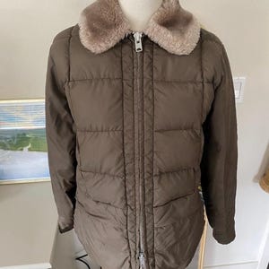 May include: A brown quilted jacket with a faux fur collar. The jacket has a full-length zipper and a tailored fit. The collar is a light brown color. The jacket is displayed on a mannequin.