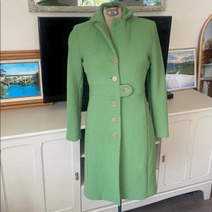 Op de afbeelding: Een lichtgroene jas met een getailleerde pasvorm, met een reverskraag, knoopsluitingen en een ceintuur in de taille. De jas wordt op een mannequin gepresenteerd, die het ontwerp en de structuur laat zien. De kleur van de jas is een levendig, lentegroen.