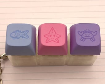 May include: A keychain featuring three square mechanical keyboard keycaps mounted on a translucent base. The left keycap is light blue with a fish icon, the center is pink with a starfish icon, and the right is purple with a crab icon.