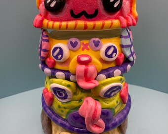 May include: A vertical stack of four colorful, sculpted clay creature faces. The top is a pink axolotl, followed by a yellow character with purple hearts, a green creature with a pink tongue, and a brown owl-like face at the base.