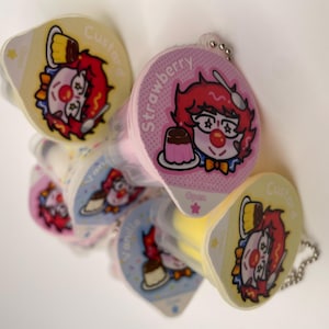 May include: A set of six colorful keychains shaped like pudding cups. Each keychain features a cartoon clown with a different flavor of pudding: strawberry, vanilla, custard, chocolate, and two others. The keychains are made of soft, squishy plastic and have a cute, whimsical design.