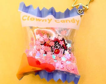 May include: A clear plastic keychain shaped like a candy bag with a gold star charm. The bag is filled with pink and white glitter and a small cartoon clown face. The bag has the words "Clowny Candy" printed on it.