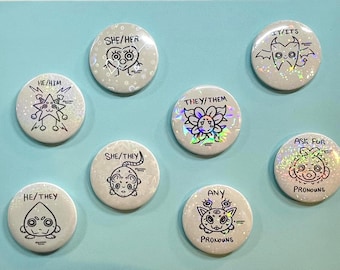 May include: A set of eight circular pin-back buttons with a holographic finish. Each button features a unique black line-art character and text indicating pronouns including HE/HIM, SHE/HER, IT/ITS, THEY/THEM, ASK FOR PRONOUNS, HE/THEY, SHE/THEY, and ANY PRONOUNS.