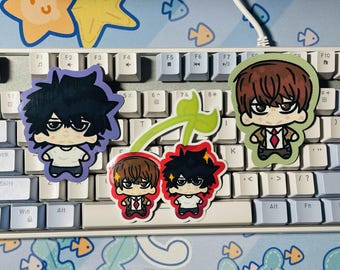 May include: Three die-cut vinyl stickers featuring chibi-style anime characters. One sticker shows a character with messy black hair, another shows a character with brown hair in a suit, and the third is a cherry-shaped design with both characters.