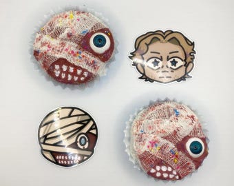 May include: Three decorated cupcakes with a white bandage and red frosting. One cupcake has a blue eye, another has a white bandage with a blue eye, and the third has a white bandage with a blue eye and a red mouth. There are two stickers, one with a cartoon face with a white bandage and blue eyes, and the other with a white bandage with a blue eye.
