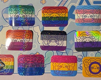 May include: A collection of nine rectangular stickers featuring a stylized creature with large eyes and circular patterns. Each sticker displays a different flag design, including various pride flags, with some featuring holographic finishes.