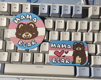 May include: Two stickers featuring a brown bear illustration and the text MAMA BEAR. One sticker is circular and the other is rectangular. Both stickers display a background with horizontal stripes in light blue, pink, and white colors.