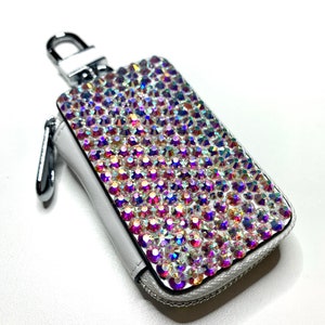 Może przedstawiać: Biały skórzany brelok do kluczy z zamkiem błyskawicznym. Przód breloka jest pokryty tęczowymi kryształami.