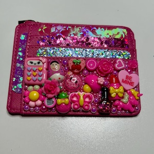 May include: A pink cardholder decorated with various pink and iridescent embellishments. The cardholder features a zip closure and is adorned with charms including a calculator, a doll, a heart that says "BE MINE", and a butterfly.