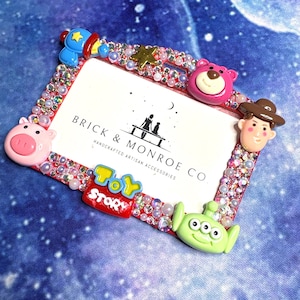 May include: A red picture frame decorated with rhinestones and Toy Story character charms. The frame features the text "TOY STORY" and "BRICK & MONROE CO." The frame is embellished with pink, white, and clear beads.