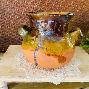 May include: A rustic brown and green ceramic pitcher with two handles. The pitcher is sitting on a white doily on a white wooden table.