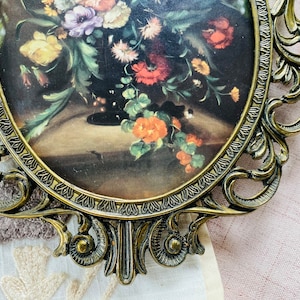 May include: A vintage oval-shaped picture frame with ornate gold metal detailing. The frame holds a floral print with a bouquet of colorful flowers in a vase.