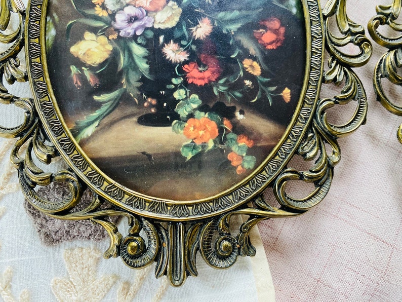May include: A vintage oval-shaped picture frame with ornate gold metal detailing. The frame holds a floral print with a bouquet of colorful flowers in a vase.