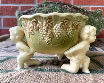 May include: A green ceramic bowl with a grapevine design is held up by two cherub figurines. The bowl has a scalloped edge and is decorated with raised grapes and leaves.