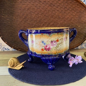 May include: A cobalt blue porcelain planter with gold accents and floral designs. The planter has three legs and is sitting on a blue mat.
