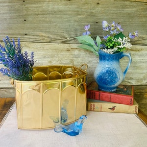 May include: A gold metal planter with a curved shape and decorative arches. The planter is filled with purple lavender flowers. A blue glass bird figurine sits on a white linen tablecloth. A blue and white pitcher with a floral design and a stack of books are in the background.