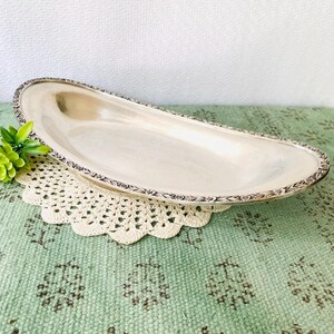 May include: A silver-colored, elongated serving tray with a decorative floral border. The tray sits on a cream-colored, crocheted doily and a green patterned surface. A small green plant is placed near the tray.
