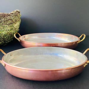 May include: Two copper pans with brass handles. The pans have a shiny, reflective interior and a reddish-brown exterior. The handles are curved and attached to the sides of the pans. A decorative basket with green foliage is in the background.