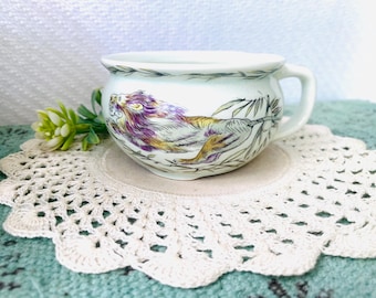 May include: White ceramic teacup with a handle, featuring a colorful illustration of a lion. The lion has purple and gold accents. The teacup sits on a beige coaster and a white crocheted doily.