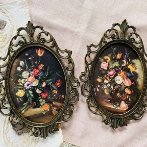 May include: Two ornate gold-toned frames with glass panels, each displaying a floral print. The frames have intricate scrollwork designs and are set against a light pink background.