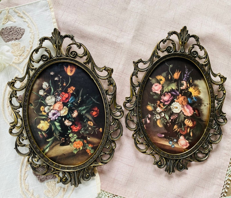 May include: Two ornate gold-toned frames with glass panels, each displaying a floral print. The frames have intricate scrollwork designs and are set against a light pink background.