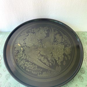 May include: A round, black serving tray with a detailed gold-colored design. The design features a pair of stylized birds amidst floral and foliage elements. The tray has a decorative border and a glossy finish, suitable for serving or display.