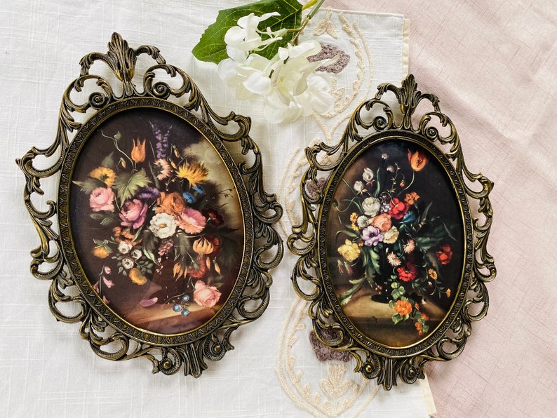 May include: Two ornate gold oval frames with intricate details, each holding a floral print with a bouquet of colorful flowers in a vase.