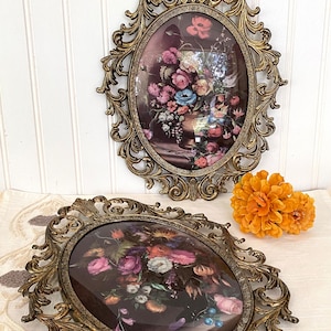 May include: Two ornate gold oval picture frames with glass fronts, each displaying a vintage floral print. The frames have intricate scrollwork and a distressed finish.