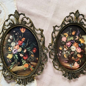 May include: Two ornate gold-toned oval frames with intricate details, each holding a vintage floral print with vibrant colors. The prints depict bouquets of roses, lilies, and other flowers.