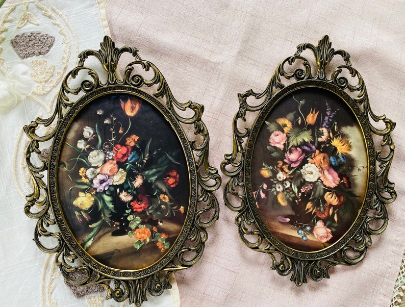 May include: Two ornate gold-toned oval frames with intricate details, each holding a vintage floral print with vibrant colors. The prints depict bouquets of roses, lilies, and other flowers.