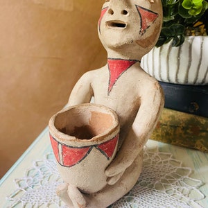 May include: A hand-painted terracotta figurine of a person with red and black geometric designs. The figurine is holding a small pot with a red and black design.