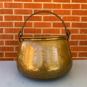 May include: A large, antique brass cauldron with a curved handle. The pot has a rounded body and a wide opening. The metal has a warm, golden hue, reflecting light. The background is a red brick wall.