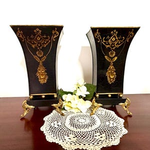 May include: A pair of black vases with a flared top and gold-colored decorative accents. Each vase features a textured surface, a rectangular base, and four ornate gold-colored metal feet. The gold detailing includes floral and leaf motifs.