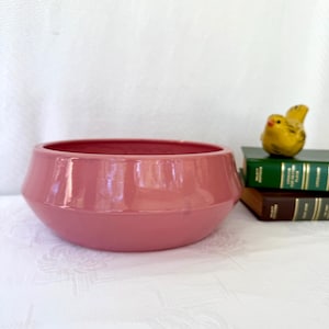 May include: A dusty rose-colored ceramic bowl with a geometric design. The bowl sits on a white surface. A small yellow bird figurine rests on a stack of green and brown books.