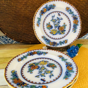 May include: Three white porcelain dessert plates with a blue and gold floral pattern. The plates have a scalloped edge and are stacked on top of each other.