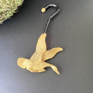 May include: A decorative brass-colored metal hook featuring a flying goose design. The hook has a curved, black metal stem with a small, gold-colored ball at the top. The goose is detailed with outstretched wings and a downward-pointing beak.