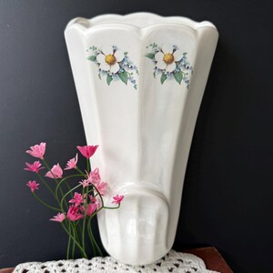 May include: A white ceramic wall vase with a scalloped top and a floral design featuring white flowers with yellow centers and blue accents. Pink flowers are placed in the vase. The vase is mounted on a dark background.